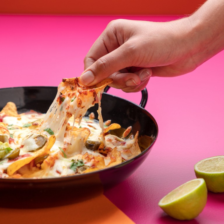 Some taking a cheese nacho from a bowl filled with cheese, nachos, salsa and guacamole